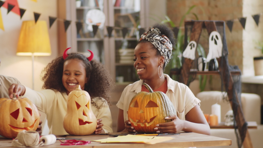 Joyous little African American family sitting at table in living room, smiling and laughing while playing with carved pumpkins on Halloween Royalty-Free Stock Footage #1060902409