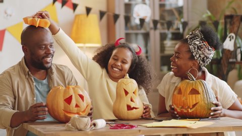 Joyous little African American family sitting at table in living room, smiling and laughing while playing with carved pumpkins on Halloween