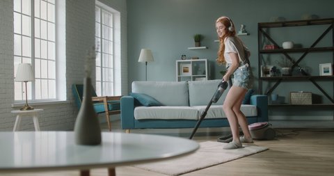 Funny girl dancing with a vacuum cleaner. Young woman cleaning her house, listening to music and expressing positive emotions - happy real people 4k footage