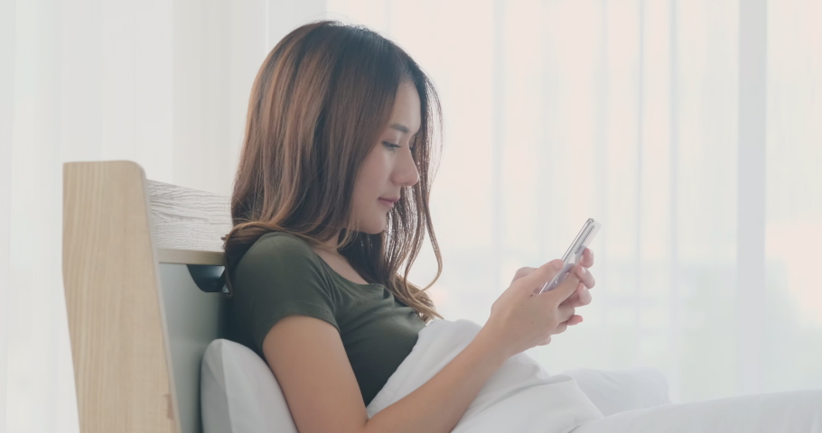 Asian young woman sit on bed with smartphone texting to boyfriend. Happy smile face of woman play smartphone and sent love message to her boyfriend. | Shutterstock HD Video #1060914004
