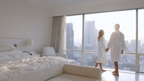 Young couple in hotel room standing by the window wearing bathrobe enjoying city views at sunset 