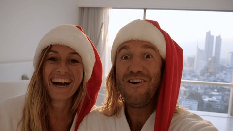 Slow motion: Young couple tasking selfies in bed with Christmas hats. Couple in hotel room in the morning taking selfies wearing Santa's red hat 