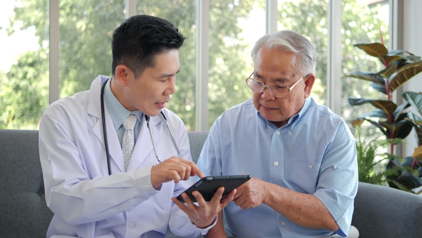 Asian man professional doctor showing medical test result explaining prescription using digital tablet app visiting senior man patient at home sitting on sofa. Elderly people healthcare tech concept.