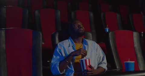 Unsatisfied spactators exept one african guy in movie theatre. People standing and walking during film in cinema while afro american guy sitting,eating popcorn and looking at screen