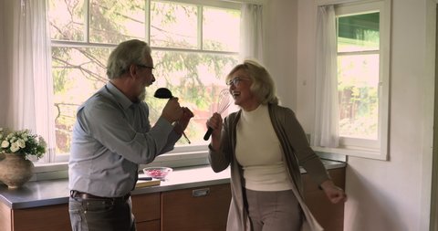 50s grey haired wife and husband hold hands while listen music sing song using kitchenware like microphones enjoy karaoke together standing in kitchen feels happy. Hobby, untroubled retirees concept