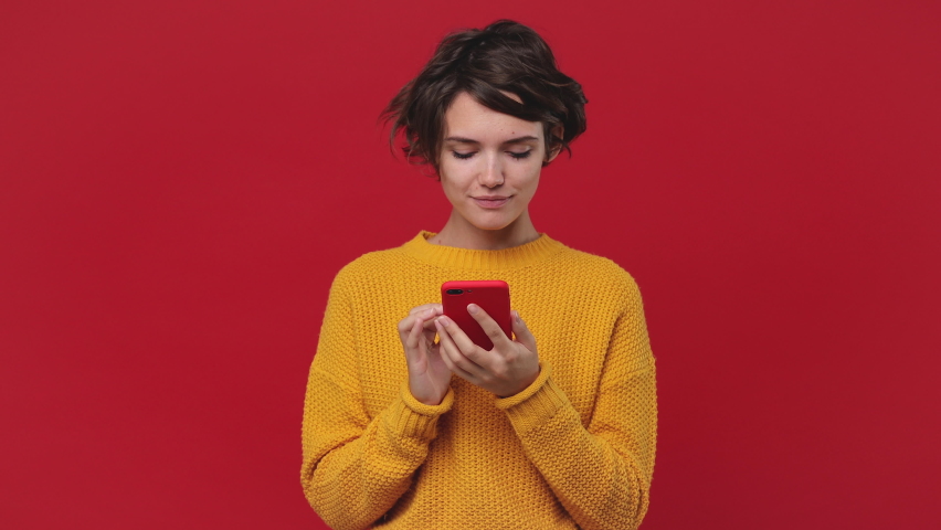 Smiling shocked happy win beautiful young woman 20s years old in yellow sweater posing isolated on red background in studio. People lifestyle concept. Look surprised wow hold using mobile cell phone | Shutterstock HD Video #1060922143
