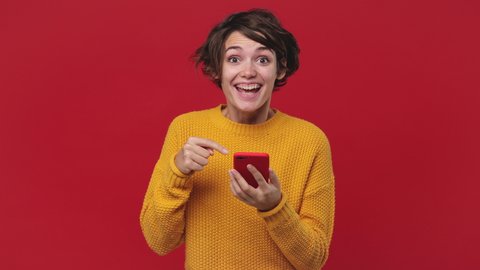 Smiling shocked happy win beautiful young woman 20s years old in yellow sweater posing isolated on red background in studio. People lifestyle concept. Look surprised wow hold using mobile cell phone