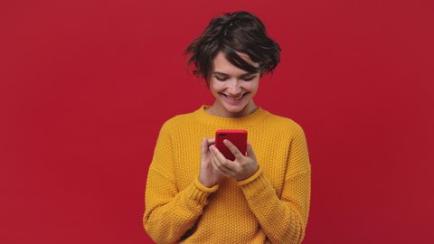 Smiling dreamful beautiful young woman girl 20s years old in yellow sweater posing isolated on red background in studio. People emotions lifestyle concept. Look surprised hold using mobile cell phone