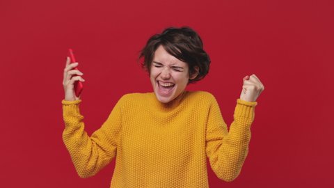Happy excited young girl 20s in yellow sweater hold talk using mobile cell phone just found out great big win news doing winner gesture isolated on red background in studio People lifestyle concept