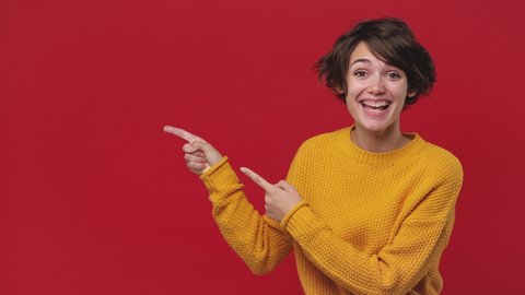 Smiling surprised beautiful young woman 20s years old in yellow sweater posing look camera pointing fingers hands on copy space workspace isolated on red background in studio. People lifestyle concept