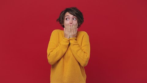 Shocked shy charming confused young woman in yellow sweater posing isolated on red background studio. People sincere emotions lifestyle concept. Looking around smile cover mouth with hands say oops