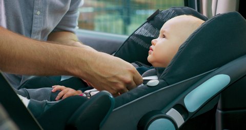 Camera inside the car. Closeup baby boy sits in the baby car seat inside of car. Young bearded father checks baby's fastened seat belts, smiles and talks to him, touching cheeks and nose. Slow motion