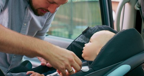 Camera inside the car. Closeup baby boy sits in the baby car seat inside of car. Young bearded father checks baby's fastened seat belts, and talks to him. Slow motion