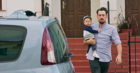 Young bearded father is carrying baby boy and puts him into the baby car seat inside of family car, while going out from the entrance stairs of his house. Slow motion