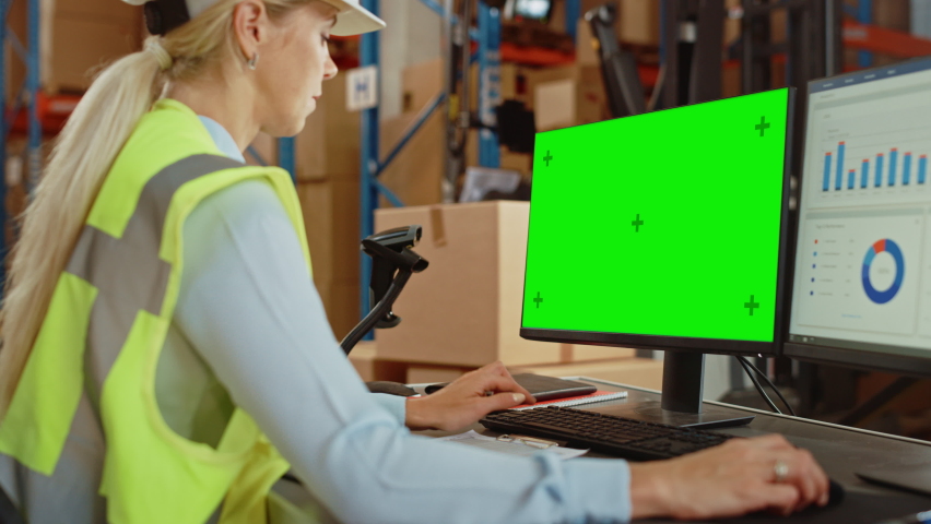 Professional Female Worker Wearing Hard Hat Uses Computer with Green Chroma Screen and Inventory Delivery Software in the Retail Warehouse full of Shelves with Goods. Distribution Logistical Center Royalty-Free Stock Footage #1060923295