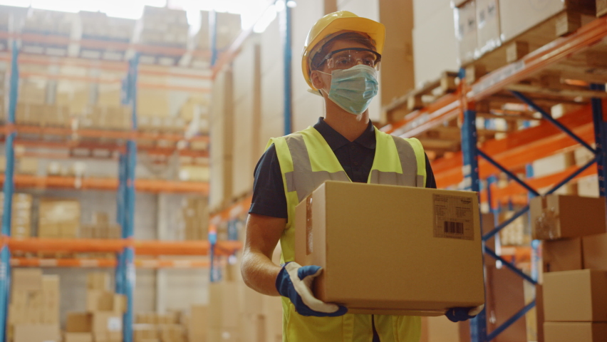 Portrait of Handsome Male Worker Wearing Medical Face Mask and Hard Hat Carries Cardboard Box Walks Through Retail Warehouse full of Shelves with Goods. Safety First Protective Workplace. Slow Motion | Shutterstock HD Video #1060923358