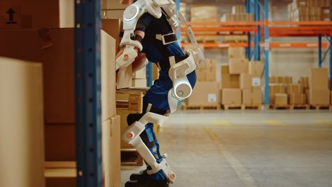 High-Tech Futuristic Warehouse: Worker Wearing Advanced Full Body Powered exoskeleton, Lifts and Walks with Heavy Pallet full of Cardboard Boxes. Delivery Exosuit amplifies Strenght. Slow Motion