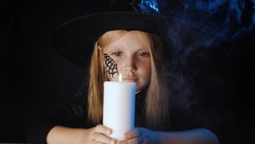 Little blonde girl in a witch hat blows out a candle in a dark room with smoke. Halloween holiday concept