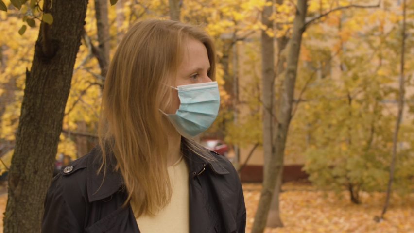 Portrait of young attractive woman in disposable protective mask standing in the autumn city. Yellow leaves on the trees, steadicam shot. Protection during viral epidemic.