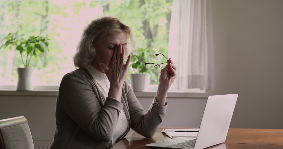 Senior woman sit at table use laptop feels tired of wireless pc usage suffers from blurry vision eyestrain problem, take off glasses relief pain. Dry eyes, aged asthenopia, age related changes concept Royalty-Free Stock Footage #1060929496