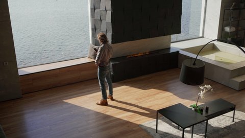 From above wide shot of young man talking on cell phone while walking towards large panoramic window in his luxury apartment, finishing call, looking at sea view and thinking with his arms crossed
