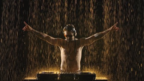 A cool bearded DJ stands behind a mixer controller, composes a new mix and lights a party in a dark studio in the rain. DJ with glasses and headphones making dance music. Close up. Slow motion.