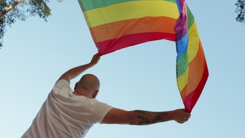 Queer man holding rainbow gay flag while parade on background of blue sky. Happy guy wearing heart sunglasses demonstrate his rights. LGBTQI, Pride Event, LGBT Pride Month, Gay Pride Symbol