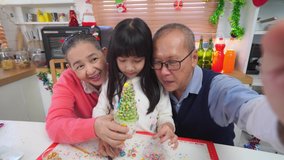 Happy Asian grandparents and cute little granddaughter taking a selfie video together, showing Christmas tree shaped cupcakes which they've just made, grandmother and granddaughter waving to video.