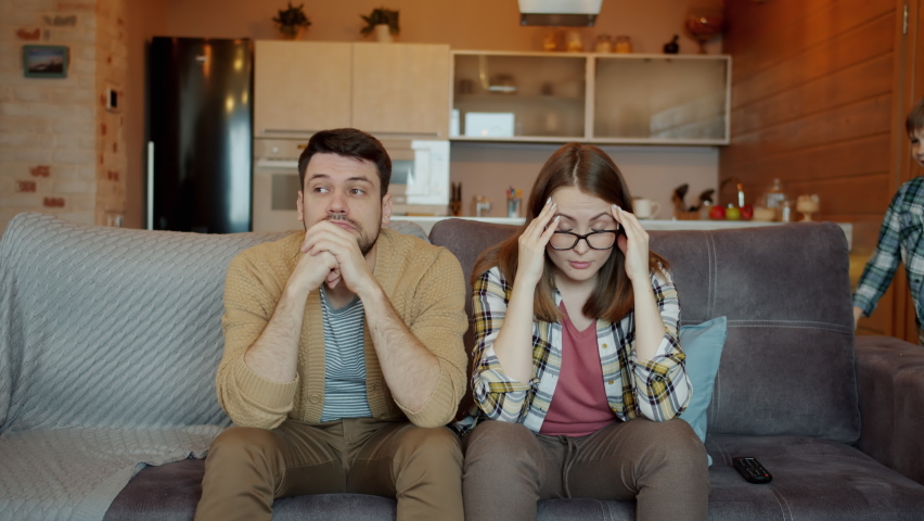 Portrait of tired parents mother and father sitting on sofa touching head while boy and girl hyperactive kids are running around fighting pillows screaming Royalty-Free Stock Footage #1060943797