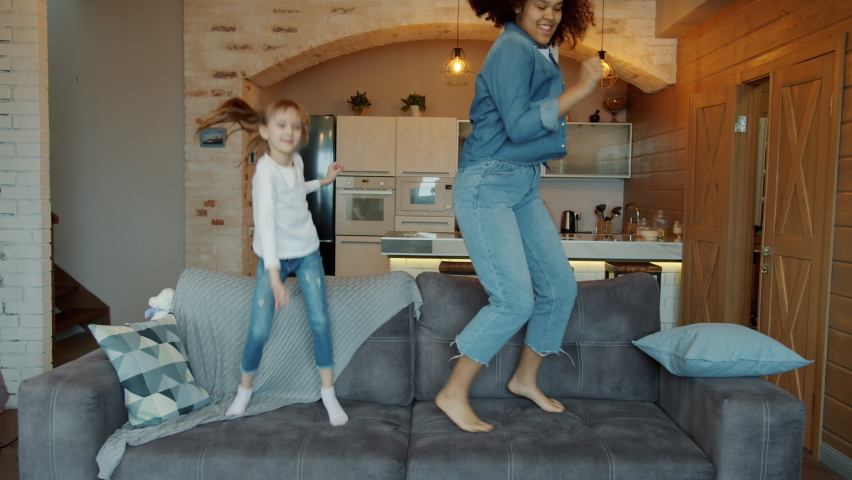 Slow motion of babysitter Afro-American woman and adorable little girl dancing jumping on sofa enjoying music at home. Lifestyle and fun concept.