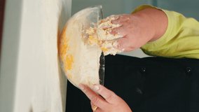 Vertical video: Chef senior woman mixing by hand bread ingredients in home kitchen in glass bowl. Retired elderly baker with bonete kneading cracked fresh eggs with wheat flour baking homemade cake