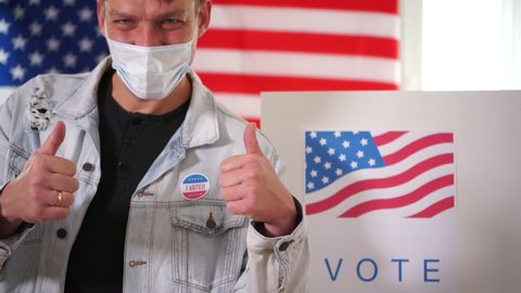 Young patriot man in mask voted in a voting booth and shows a thumbs up sign. Sticker vote on the jacket. US elections 2020 concept