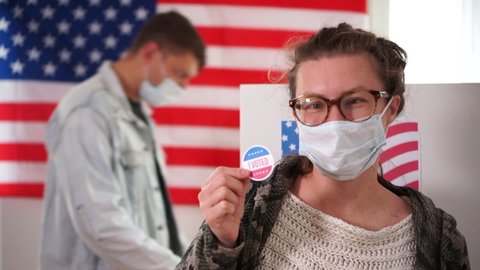 Woman in medical mask showing I voted by mail sticker with US flag as background. In the background, a man approaches a voting booth, us elections 2020, covid-19 coronavirus pandemic