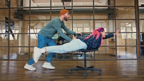Office superhero. Young man riding his colleague woman with pink hair on chair pretending she is flying, side view