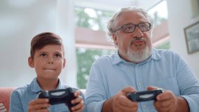 Portrait of cheerful grandson and grandfather playing video game in living room. Happy boy and senior man having fun with joysticks at home. Smiling grandparent and child using gamepads for video game