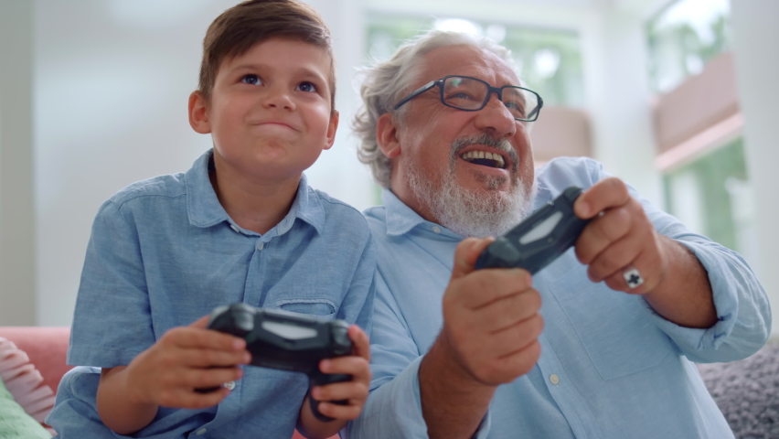 Portrait of cheerful grandson and grandfather playing video game in living room. Happy boy and senior man having fun with joysticks at home. Smiling grandparent and child using gamepads for video game | Shutterstock HD Video #1060947505
