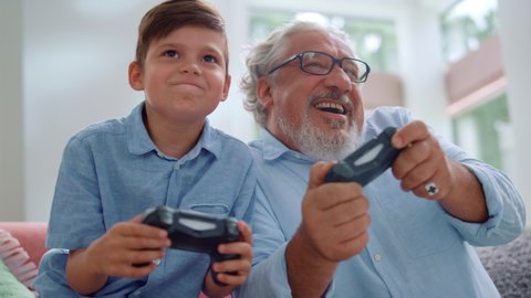 Portrait of cheerful grandson and grandfather playing video game in living room. Happy boy and senior man having fun with joysticks at home. Smiling grandparent and child using gamepads for video game