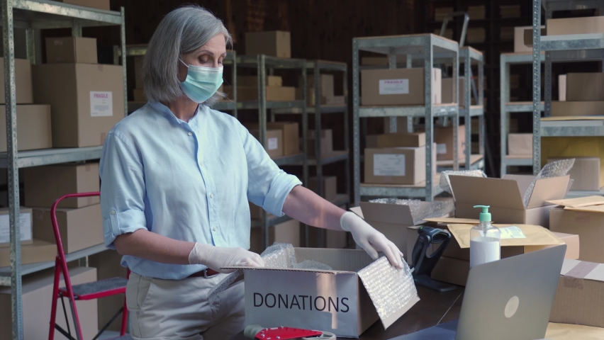 Mature female warehouse worker volunteer wearing face mask working in shipping delivery charitable stock organization packing donations box. Covid 19 coronavirus donating and volunteering concept. Royalty-Free Stock Footage #1060958170