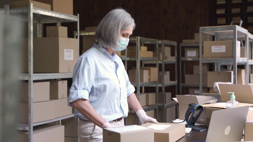 Female manager supervisor wearing face mask preparing fast drop shipping delivery boxes, putting packages on shelves while male courier taking ecommerce orders to deliver from warehouse storage. Royalty-Free Stock Footage #1060958173
