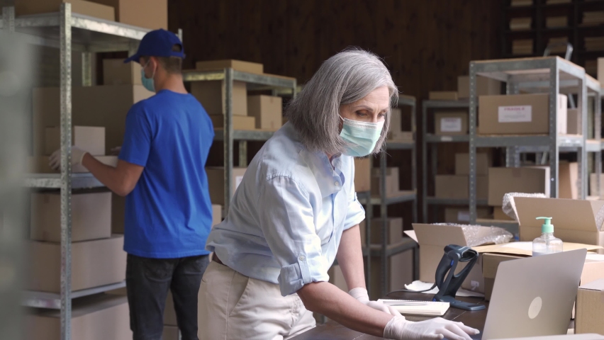 Female manager supervisor wearing face mask preparing fast drop shipping delivery boxes, putting packages on shelves while male courier taking ecommerce orders to deliver from warehouse storage. | Shutterstock HD Video #1060958173