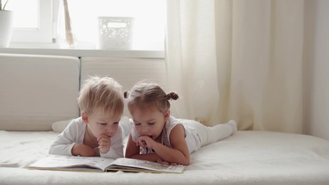 Friendship, family, education concepts  - Two smiling preshool, toddler children read large interesting book of fairy tales on bed. Siblings little twins have fun, happy kids on quarantine at home