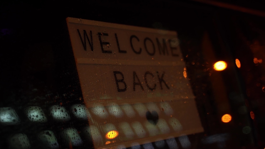 Lightbox with a sign WELCOME BACK behind a glass door of the cafe during the rain at night. We're open again after quarantine. Please wear a face mask and keep your distance Royalty-Free Stock Footage #1060959913