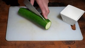 This video shows anonymous hands cutting, chopping, and slicing a fresh cucumber on a kitchen cutting board with a chef's knife.