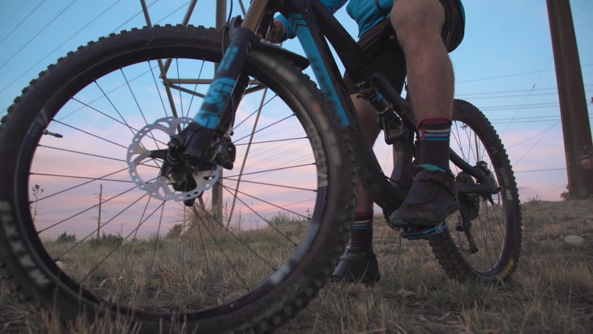 Medium-wide crane shot of a male mountain biker, atop a hill underneath a power line tower as the low-sun creates a dramatic sky. He stares and ponders with hopeful inspiration.