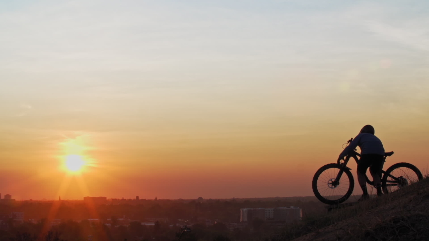 A male mountain biker, overlooking a town skyline on a hill, hoists his bike over his head as the low sun silhouettes his form.
