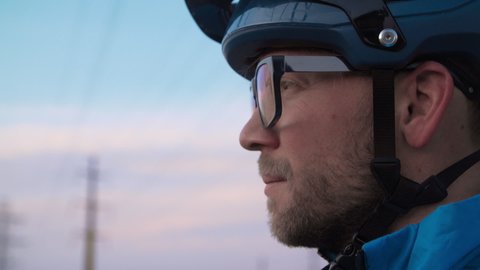 Close up shot of a male mountain biker, atop a hill observing a town skyline as the low-sun creates a dramatic sky and serene setting. He stares and ponders with hopeful inspiration.