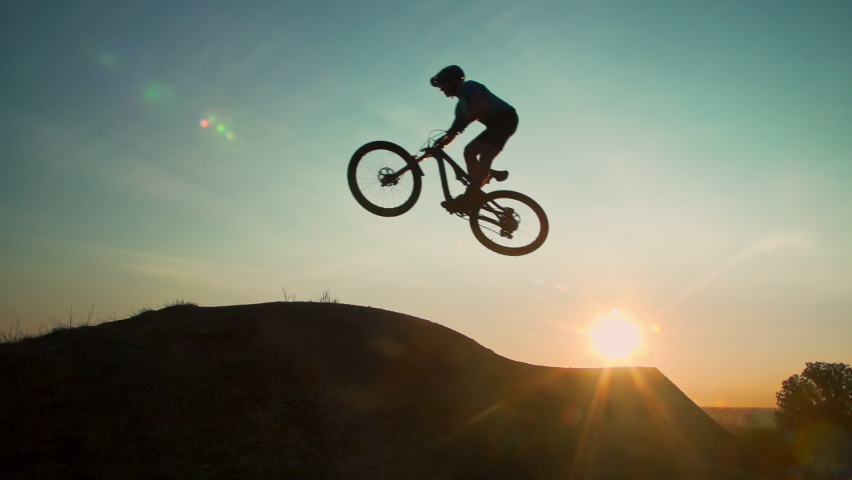 Man on mountain bike launches over a dirt ramp in a bike park into the sky. His silhouette passes in front of the sun. Shot in glorious slow motion at 240 fps. Royalty-Free Stock Footage #1060965193
