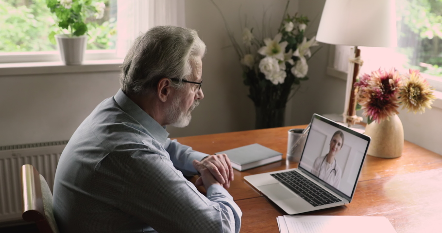 Side view hoary old man in glasses sit at table at home look at laptop screen answer question about health talks with therapist by videoconference call application. Remote medical consultation concept Royalty-Free Stock Footage #1060968217