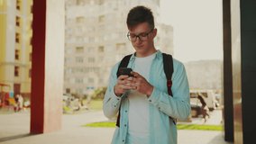 Student walking with smartphone. Video portrait of young man wearing glasses walking with smartphone at the sunset. He reading something and looking straight. Outdoor, gadget using concept