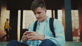 Student sitting on the stairs with smartphone, smiles and text with friends. Close up video portrait of young man with blonde hair in casual clothes sitting on the stairs, smiles and using smartphone